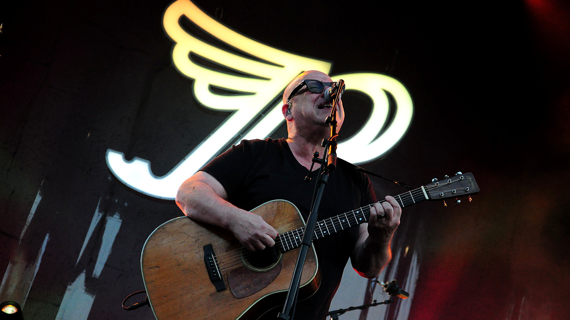 Pixies-Sänger Black Francis singt mit Akustikgitarre vor dem großen beleuchteten Bandlogo