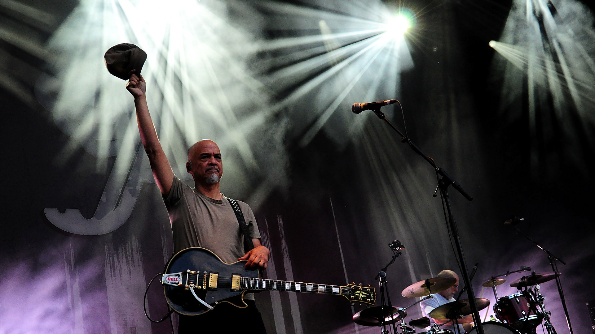 Pixies-Gitarrist Joey Santiago hebt seine Kappe unter weißen Lichtstrahlen auf der Bühne