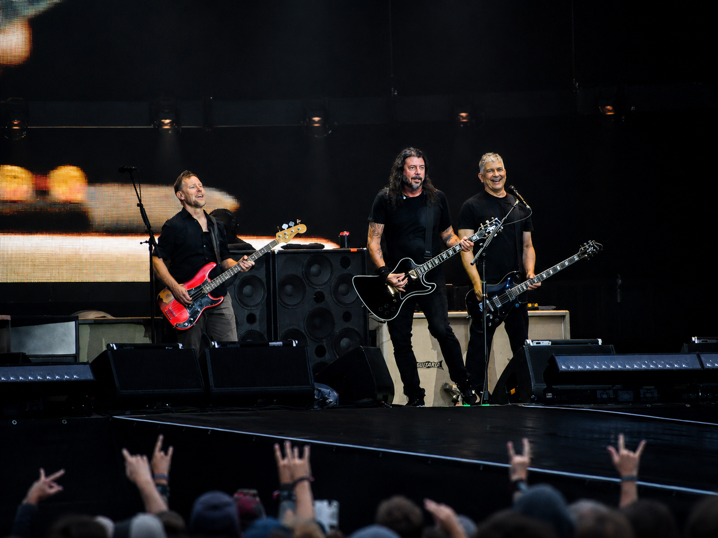 Drei Musiker der Foo Fighters stehen lachend mit Gitarren und rotem Bass gemeinsam auf der Bühne.