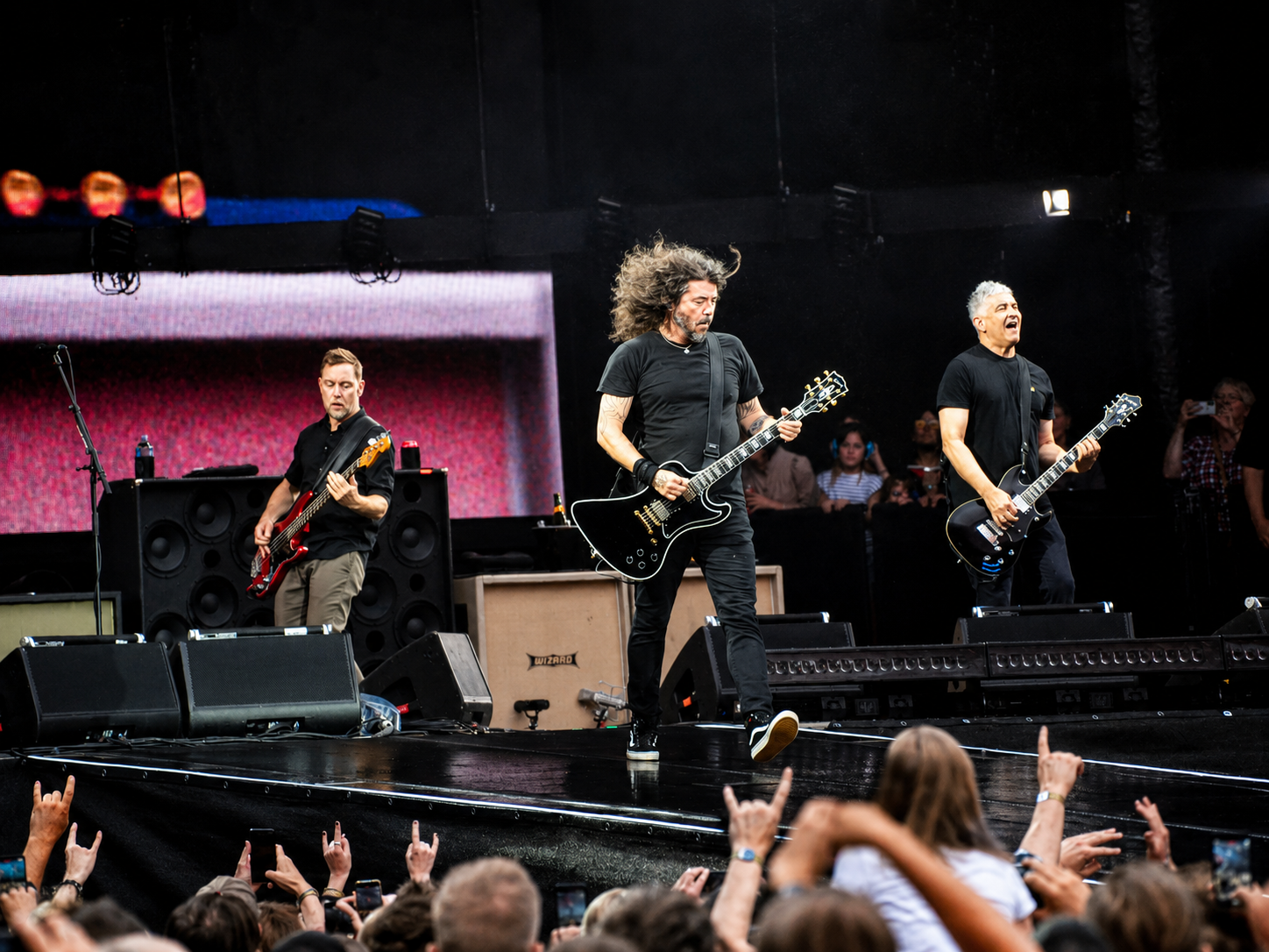 Drei Musiker der Foo Fighters spielen gemeinsam auf dem Bühnensteg vor jubelnden Fans im Olympiastadion Berlin.