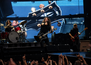 Foo Fighters live im Olympiastadion Berlin mit Sänger, Schlagzeuger und Bassist vor einer großen Gitarrenprojektion.