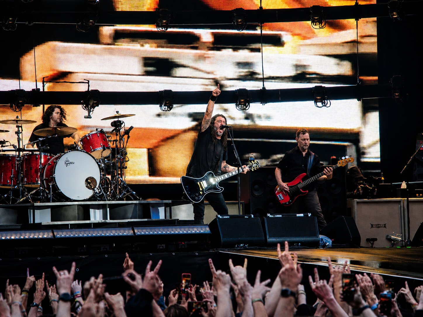 Frontmann der Foo Fighters steht mit erhobenem Arm vor Schlagzeuger und Bassist auf der Berliner Konzertbühne.