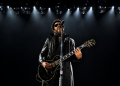 Lenny Kravitz mit schwarzer Gitarre und Sonnenbrille bei einem Konzert unter hellem Bühnenlicht