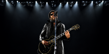 Lenny Kravitz mit schwarzer Gitarre und Sonnenbrille bei einem Konzert unter hellem Bühnenlicht