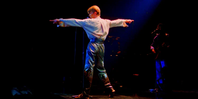 David Bowie steht mit ausgestreckten Armen auf einer dunkel beleuchteten Konzertbühne – Pressefoto zur Show STAGE in Berlin 2027