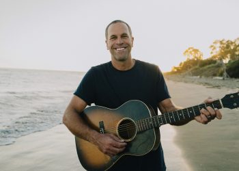 Jack Johnson steht mit einer Akustikgitarre am Strand vor dem Meer und lächelt in die Kamera.
