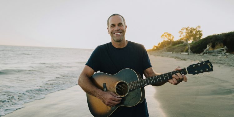 Jack Johnson steht mit einer Akustikgitarre am Strand vor dem Meer und lächelt in die Kamera.