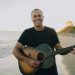 Jack Johnson steht mit einer Akustikgitarre am Strand vor dem Meer und lächelt in die Kamera.