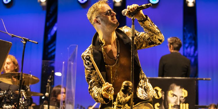 Philipp Hochmair mit Mikrofon, Sonnenbrille und goldener Jacke bei „Jedermann Reloaded“ auf der Bühne.