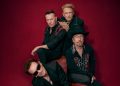 Die vier Mitglieder von U2 auf einem Pressefoto vor dunkelrotem Hintergrund zur Welttournee 2027 mit Konzert im Olympiastadion Berlin.